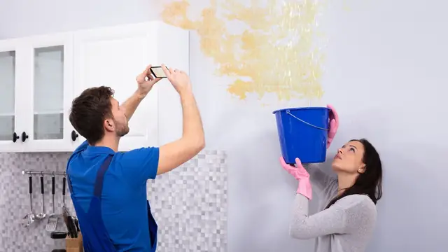 Man photographing a ceiling water stain while a woman holds a bucket under the leak