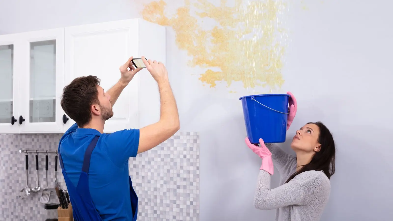 Man photographing a ceiling water stain while a woman holds a bucket under the leak