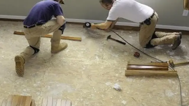 Two workers kneeling on a bare subfloor with hand tools and trim boards